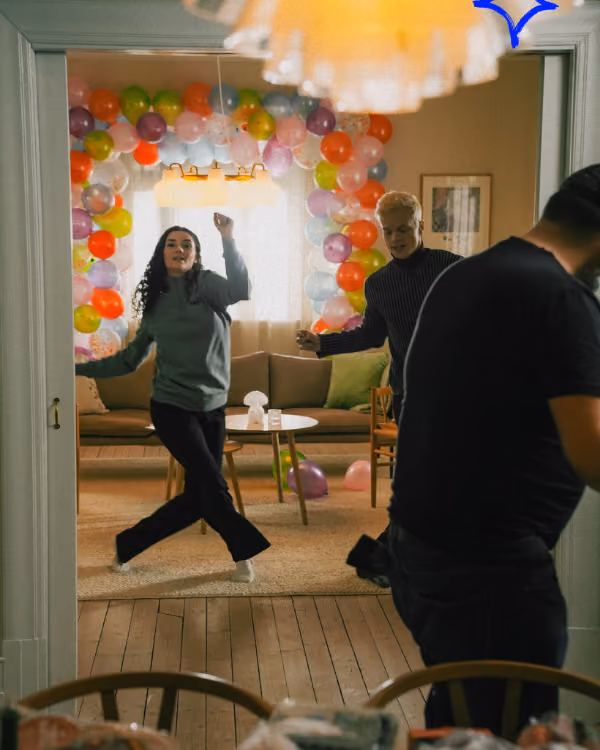 To personer danser i et rom pyntet med fargerike ballonger, mens en annen person står i forgrunnen.