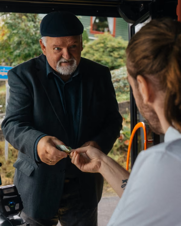 Mann med sort lue betaler bussjåfør med kontanter gjennom bussvindu.