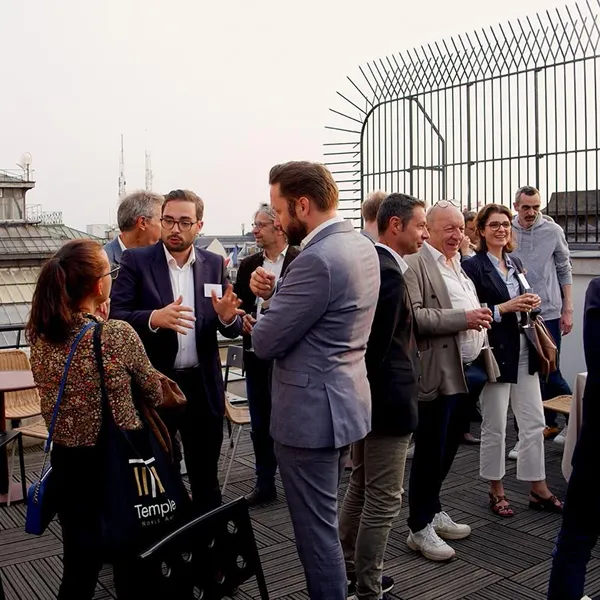 Groupe de personnes habillées professionnellement discutant lors d'un événement en plein air sur une terrasse avec une vue urbaine en arrière-plan.