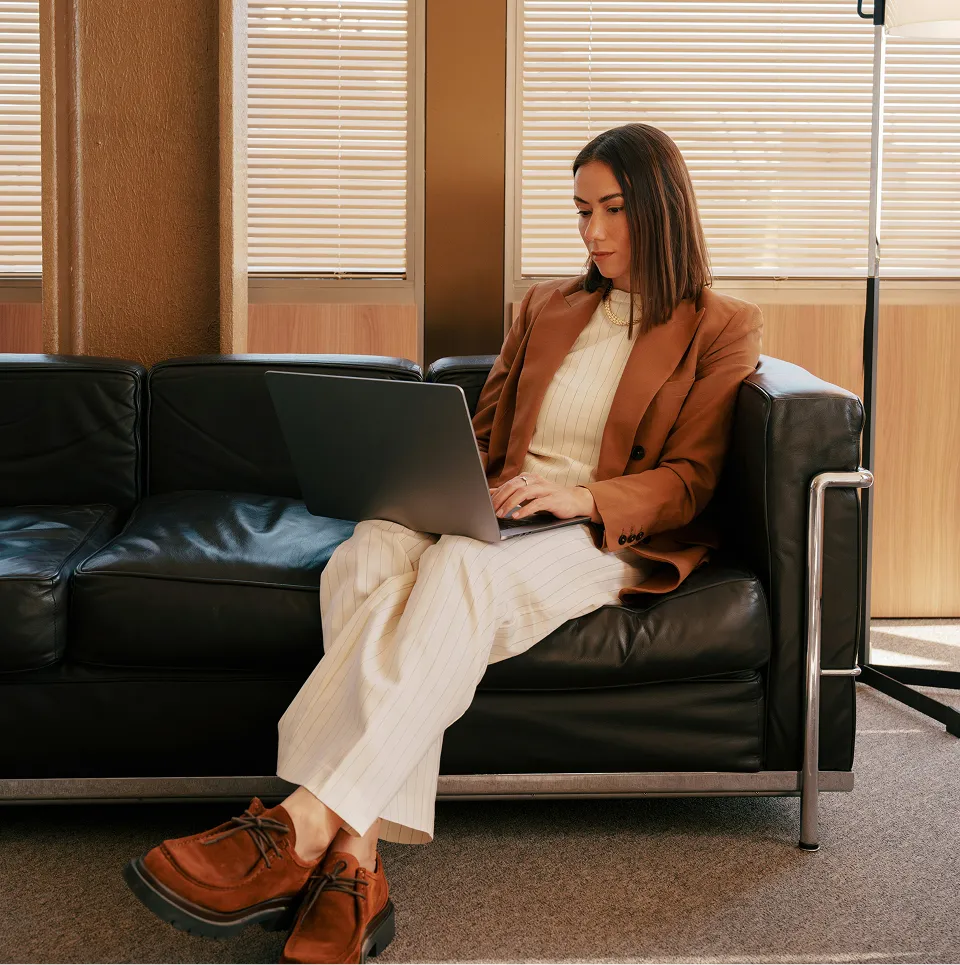 Femme assise sur un canapé en cuir noir, travaillant sur un ordinateur portable dans un bureau lumineux.