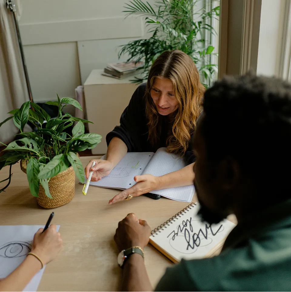 Trois personnes collaborent autour d'une table avec des cahiers et une plante verte.
