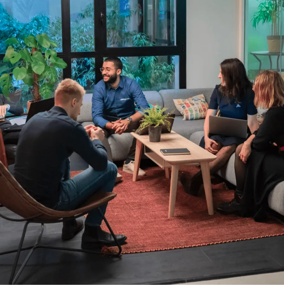 Un groupe de cinq personnes discutant dans un espace de travail moderne avec canapé gris, table basse en bois et plantes vertes.