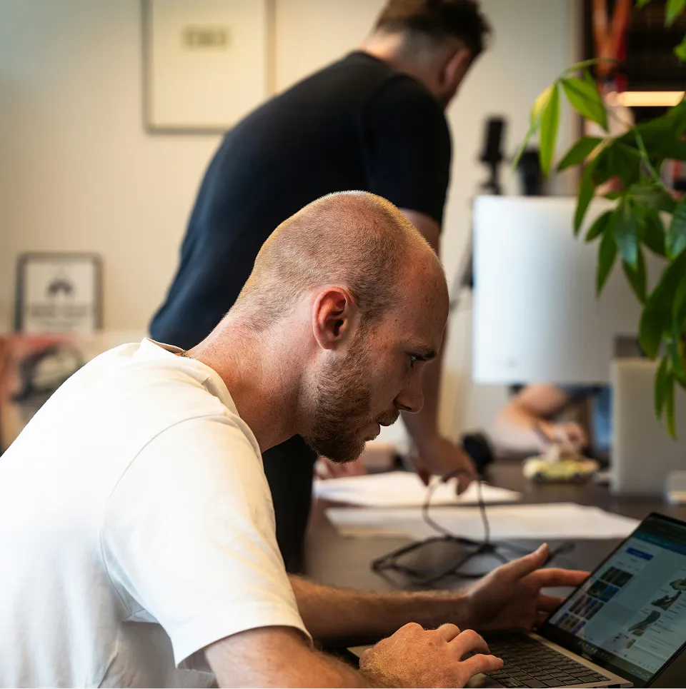 Un homme chauve portant un t-shirt blanc travaille sur un ordinateur portable dans un bureau avec une autre personne en arrière-plan.