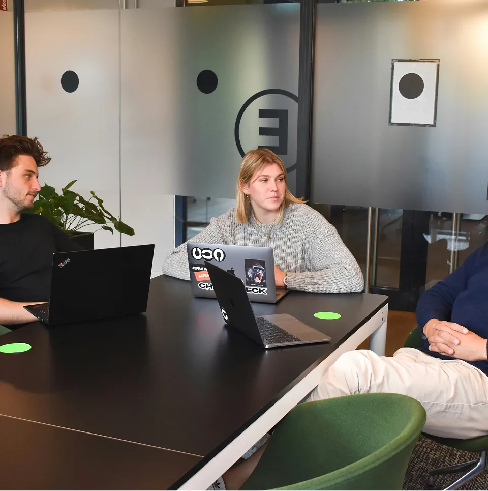 Trois jeunes adultes assis autour d'une table noire dans une salle de réunion moderne, utilisant des ordinateurs portables.
