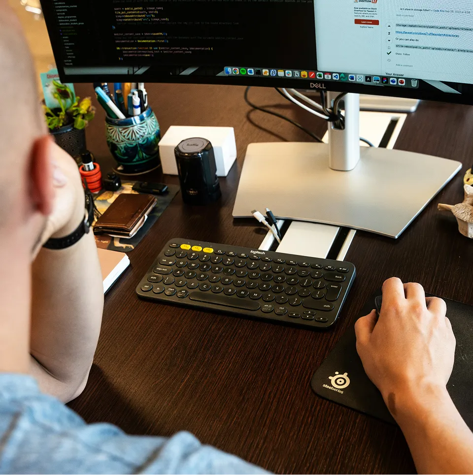 Une personne utilisant une souris sur un tapis SteelSeries devant un clavier Logitech et un grand écran d'ordinateur affichant du code.