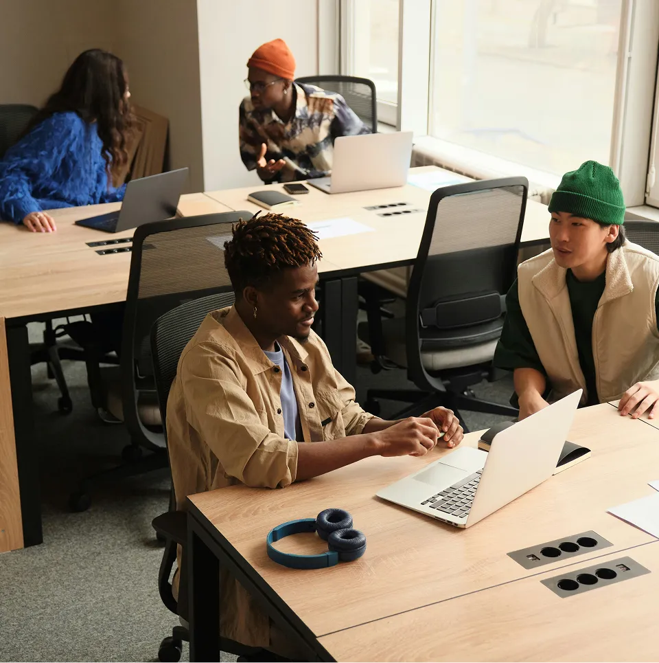 Quatre jeunes adultes discutant et travaillant ensemble dans un bureau lumineux avec des ordinateurs portables sur des bureaux en bois.
