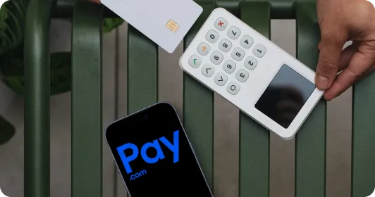 Hand holding a card reader near a credit card beside a phone showing a payment app screen.