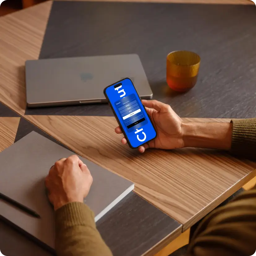 Person holding smartphone showing login screen, with closed laptop, notebook, and cup on wooden table.