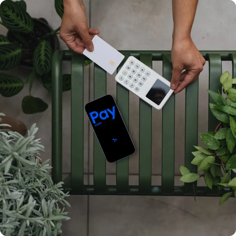 Hands holding a white card and payment device near a phone showing Pay.com on a green bench.