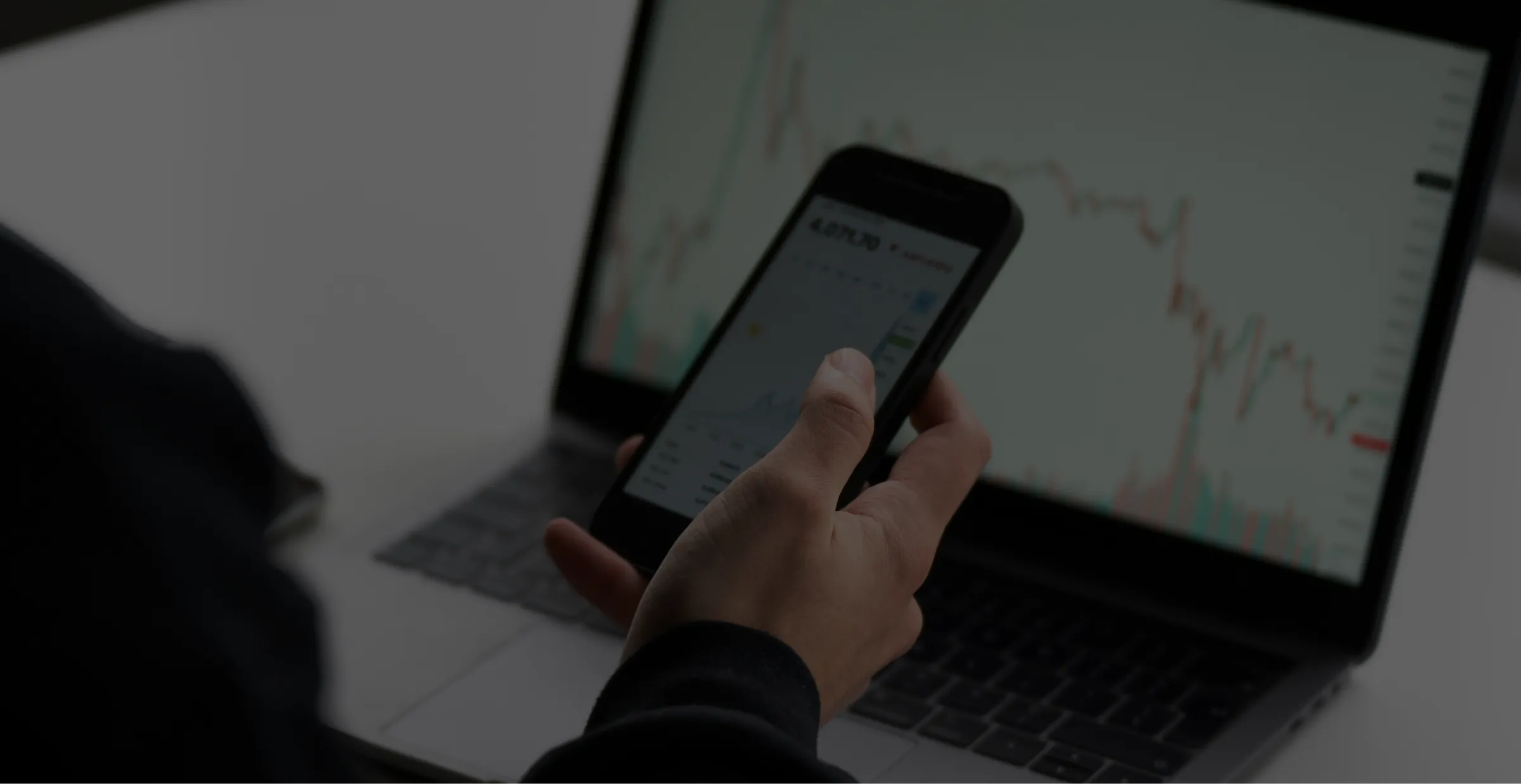 Person holding smartphone showing financial data, laptop screen in background with stock charts.