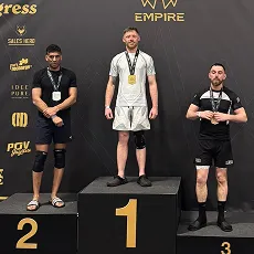 Three men stand on a winners' podium wearing medals at a powerlifting event with black background.