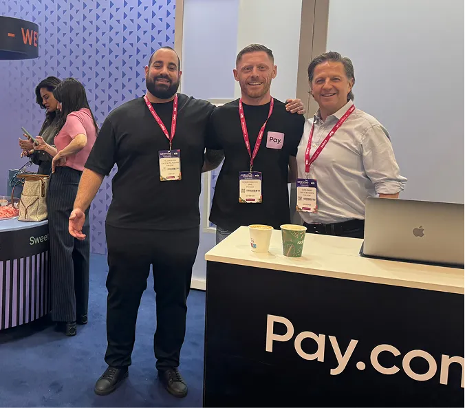 Three men with conference badges standing together smiling near a Pay.com booth with coffee cups on the counter.
