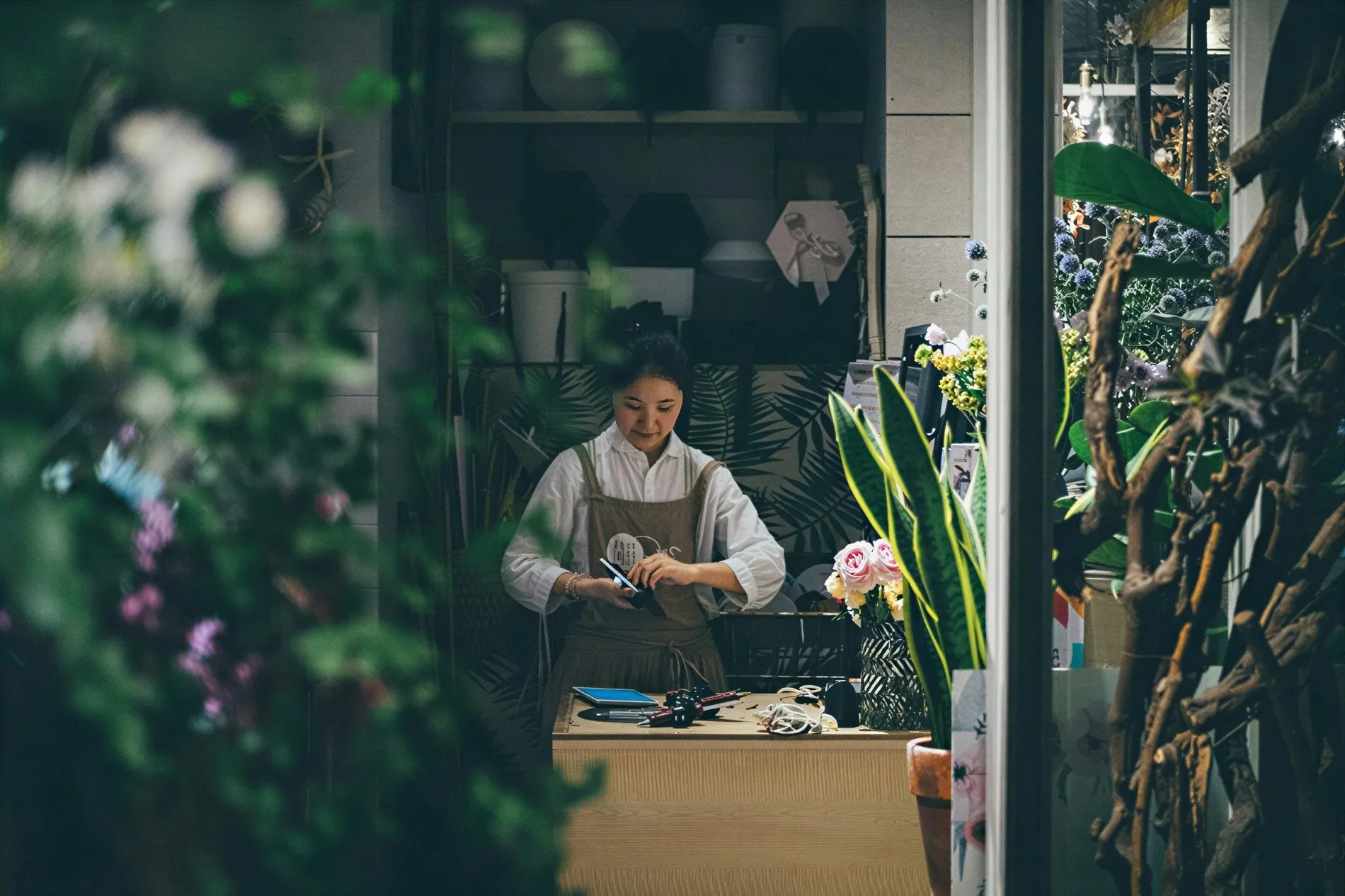 A woman wearing an apron arranging flowers behind a wooden counter in a flower shop filled with plants and floral decorations.