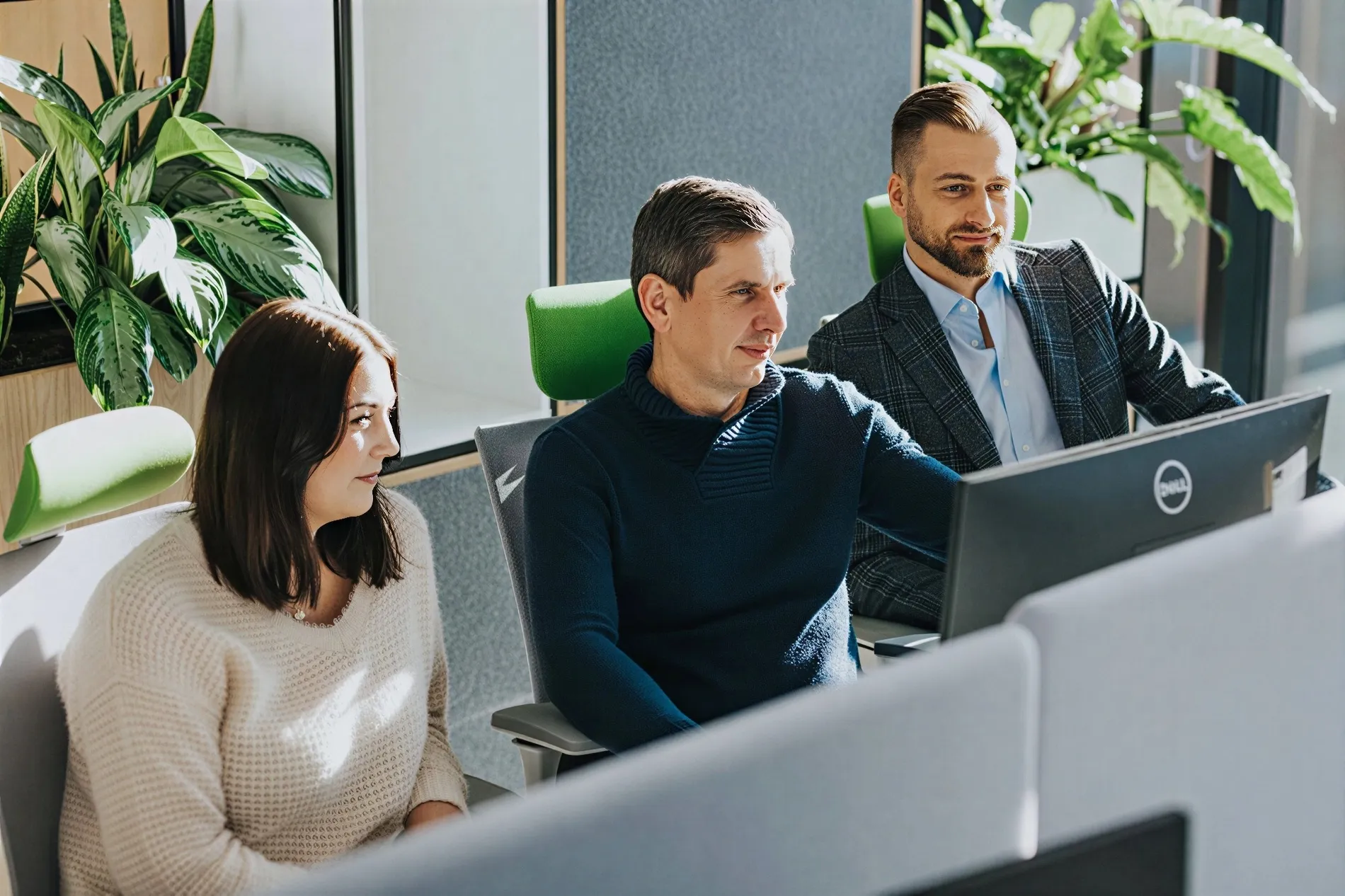 Three colleagues sitting in an office with green chairs, focusing on a computer screen.