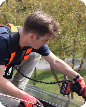 Young man wearing red gloves and an orange safety harness working on a rooftop installing solar, using a cordless power drill.