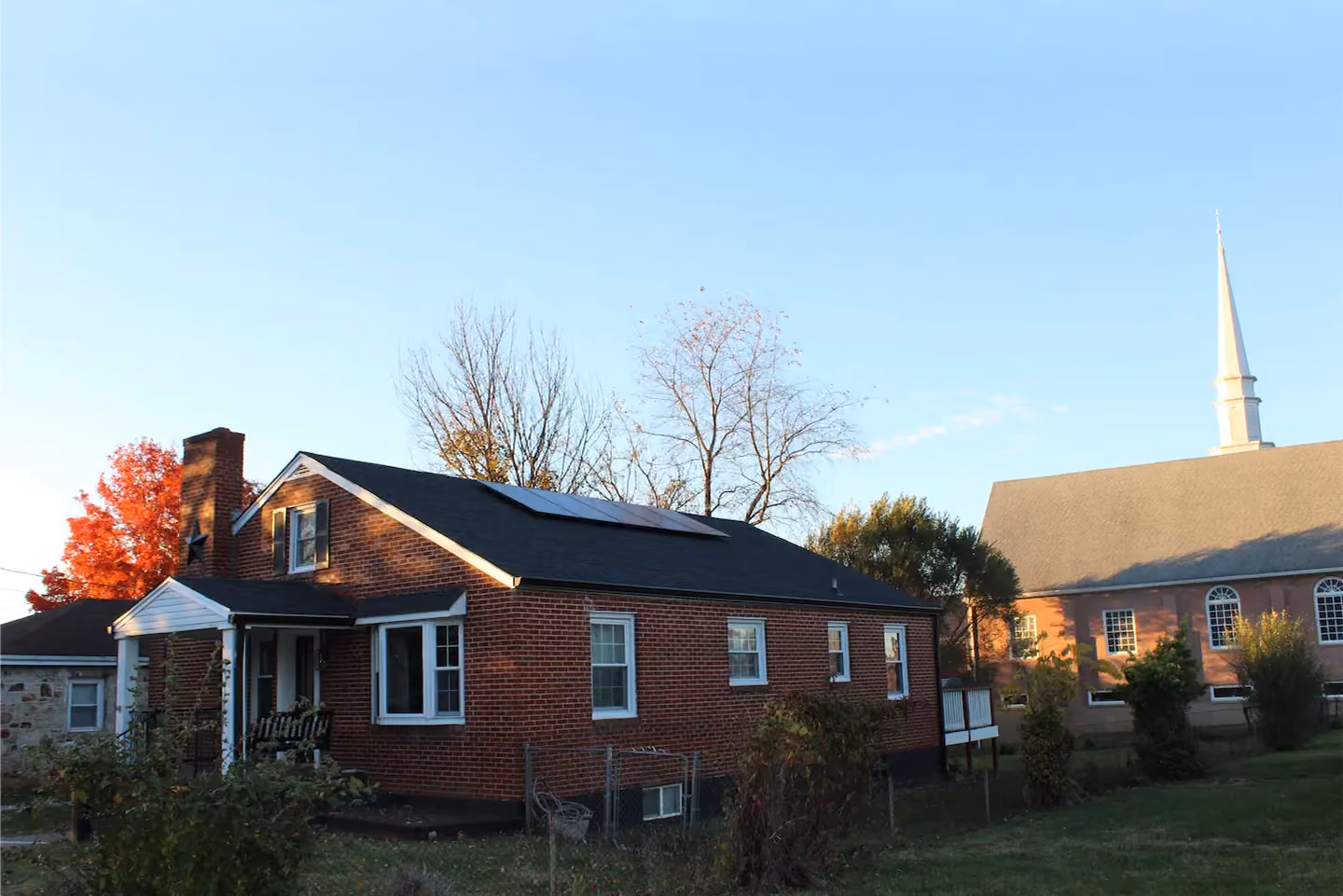 Brick house with solar panels on the roof beside a large church with a tall white steeple under a clear blue sky.