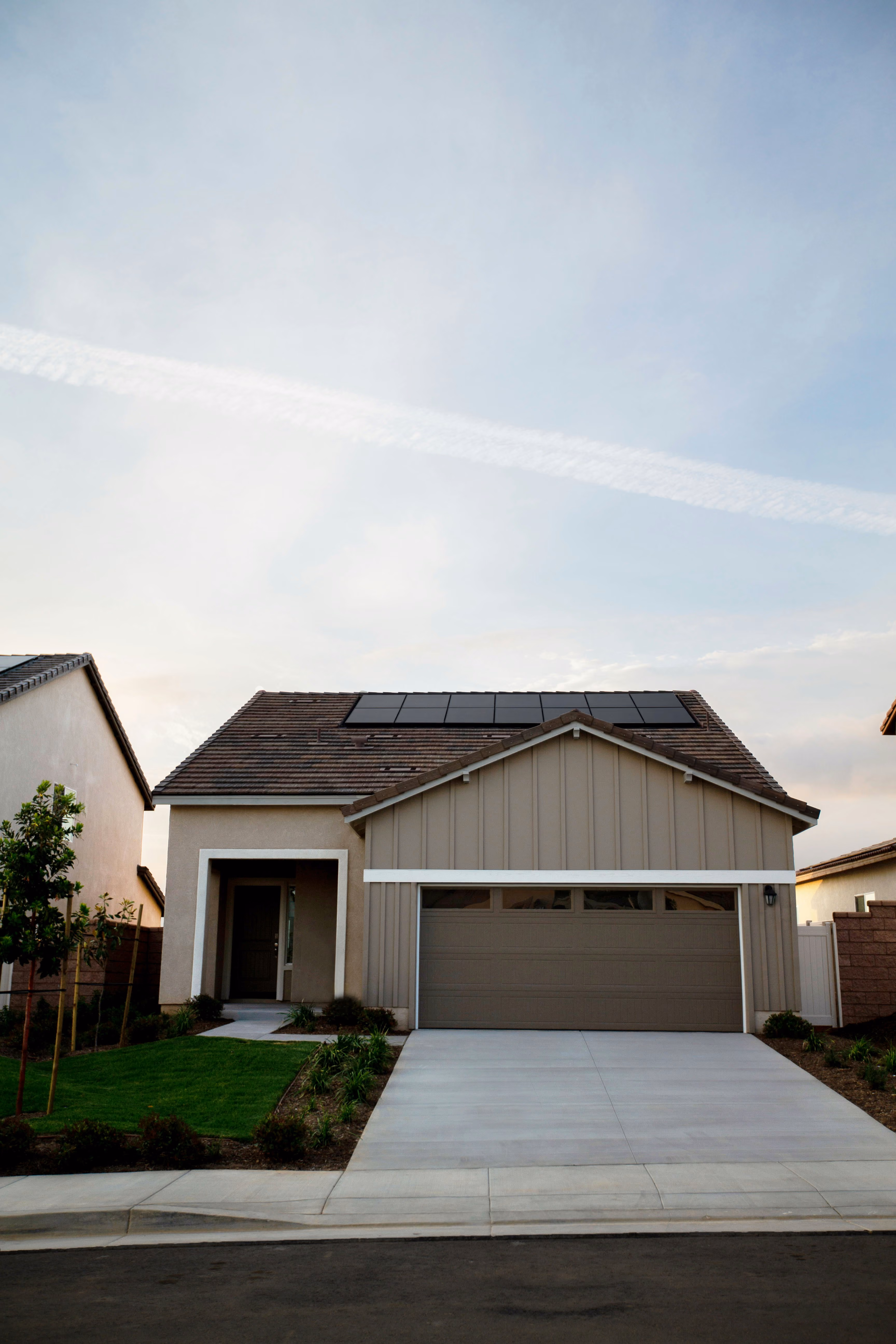 Modern single-story house with beige siding, a double garage door, landscaped front yard, and solar panels on the roof under a partly cloudy sky.