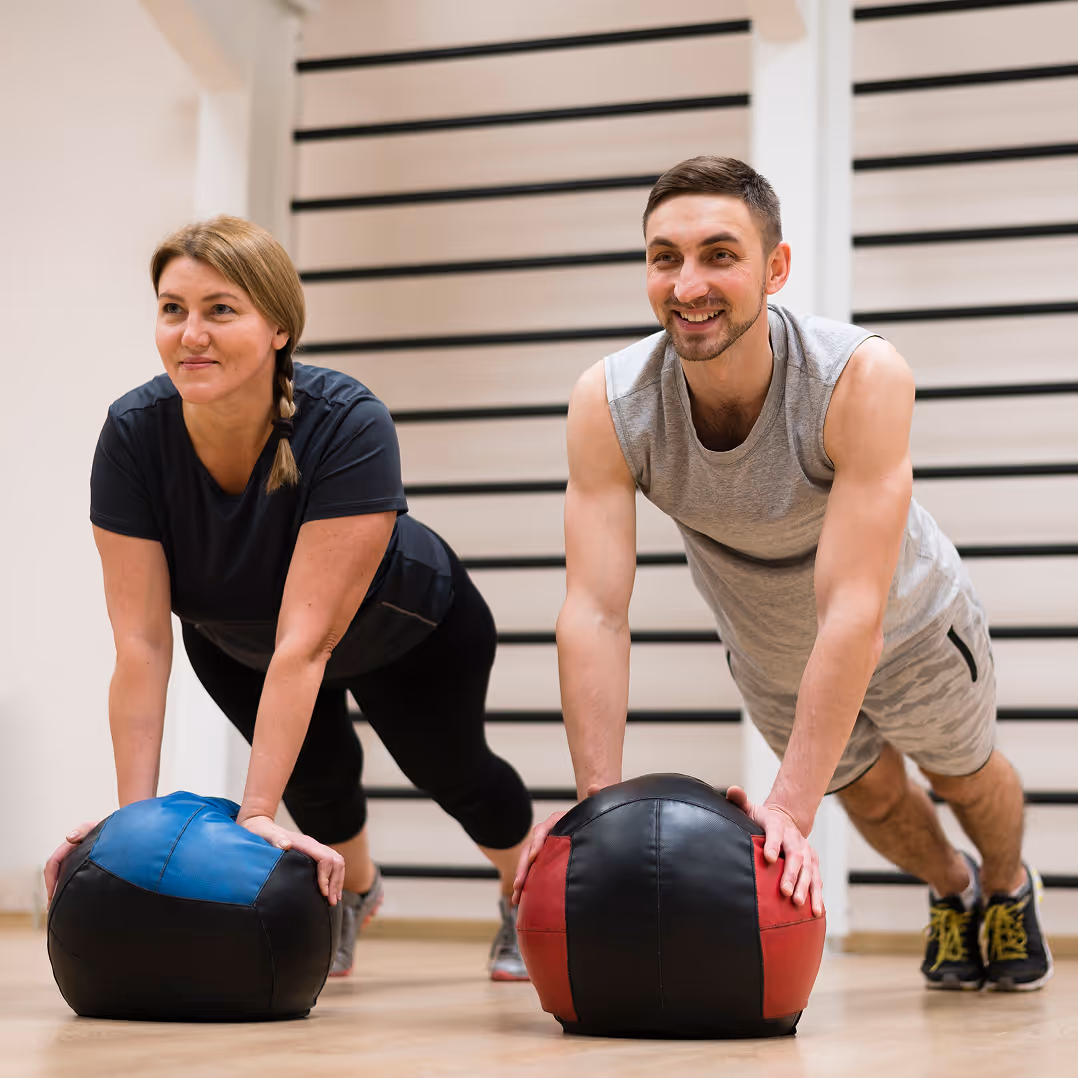 Un homme et une femme font des pompes en appui sur des ballons médicinaux en salle de sport.