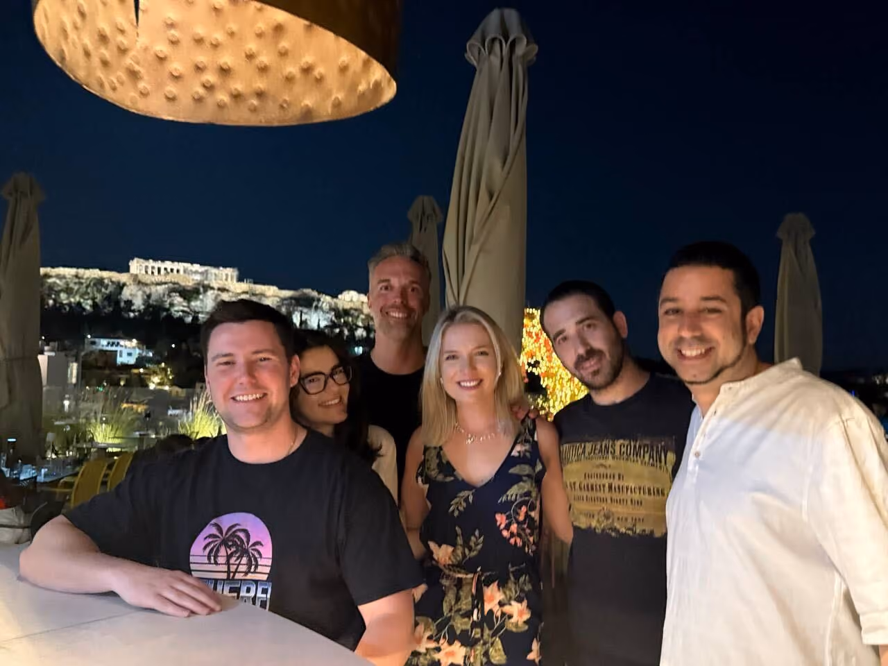 Group of six smiling people posing outdoors at night with illuminated ancient ruins in background.