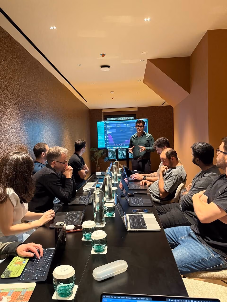 Group of people in a meeting room watching a presenter explaining data on a screen with laptops open.