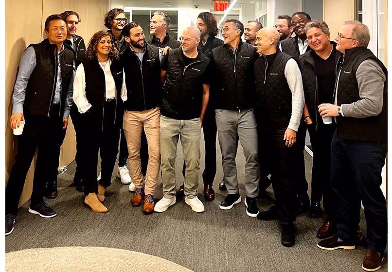 Group of twelve people in black quilted vests smiling and standing together in an office hallway.