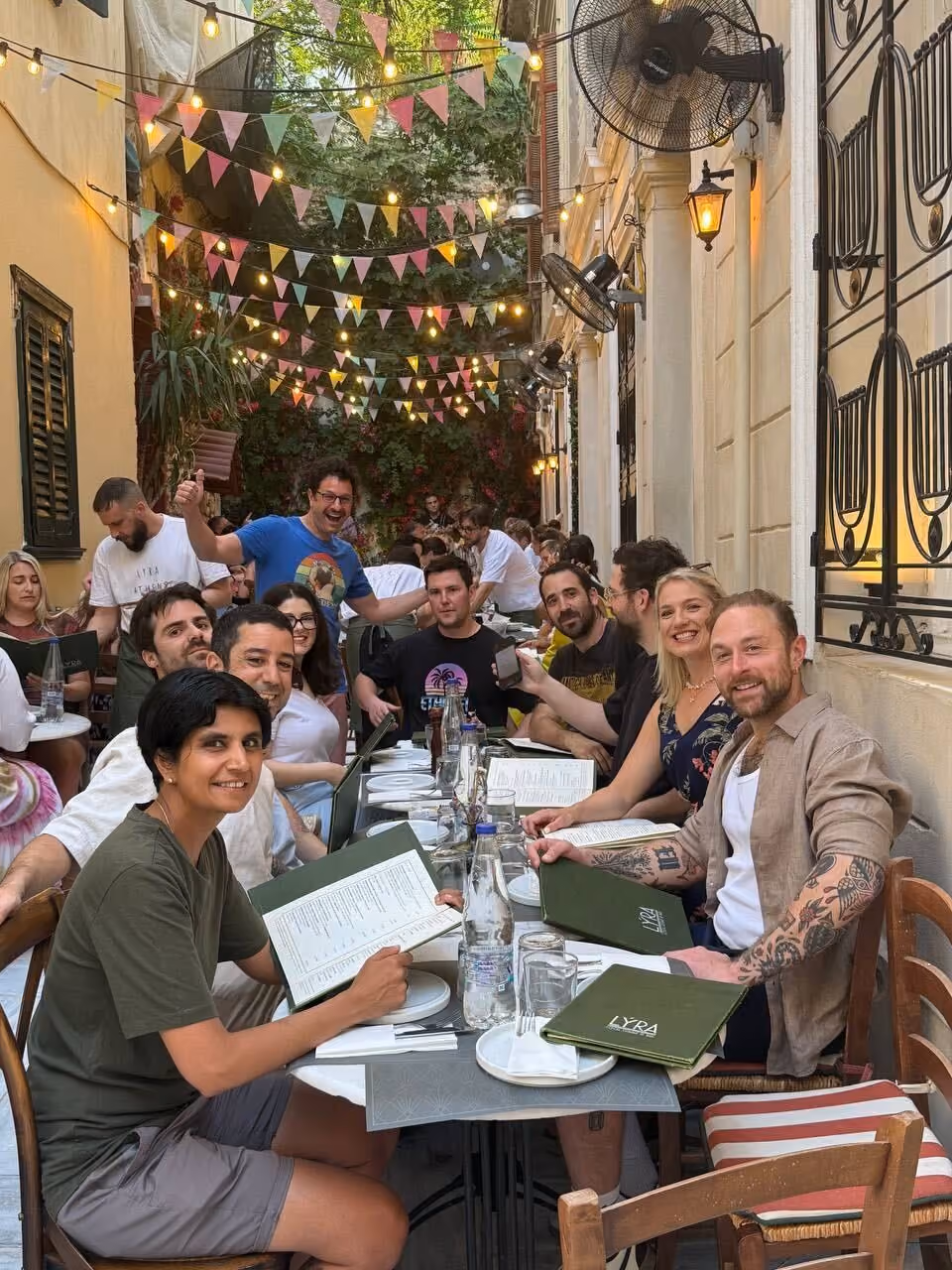 Group of people sitting at a long table outside a restaurant with menus and string lights overhead.