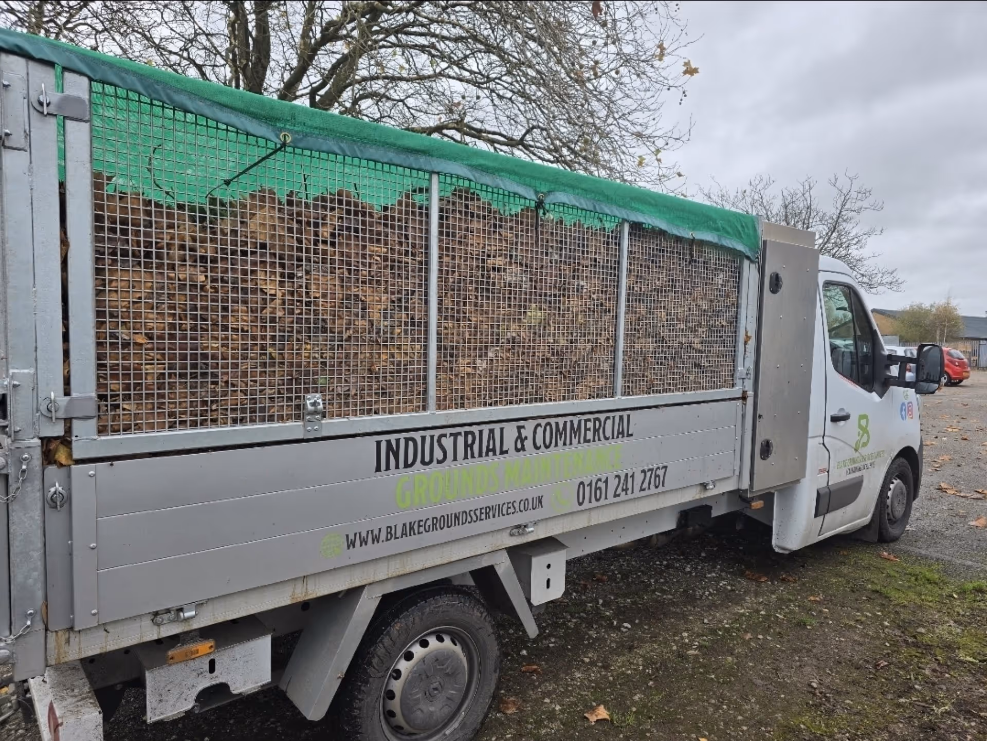 White truck loaded with logs and covered by green netting for industrial grounds maintenance.