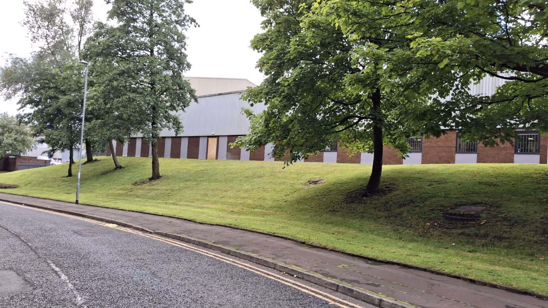 Road with sidewalk next to a grassy hill with trees and a brick and metal industrial building.