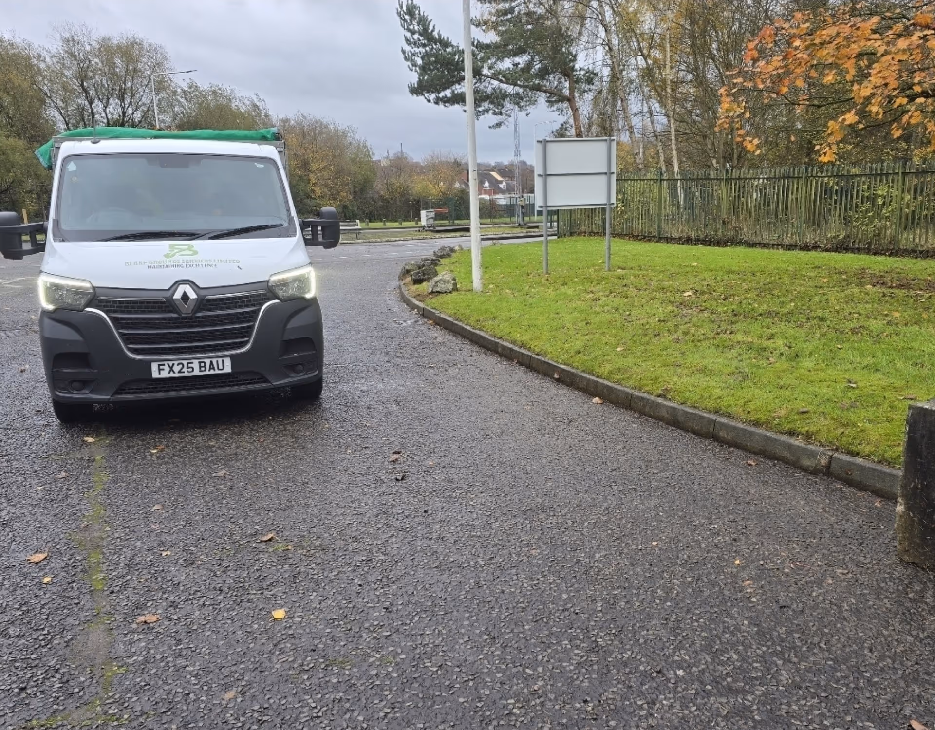 White Renault van with headlights on parked on asphalt near green grass and autumn trees.