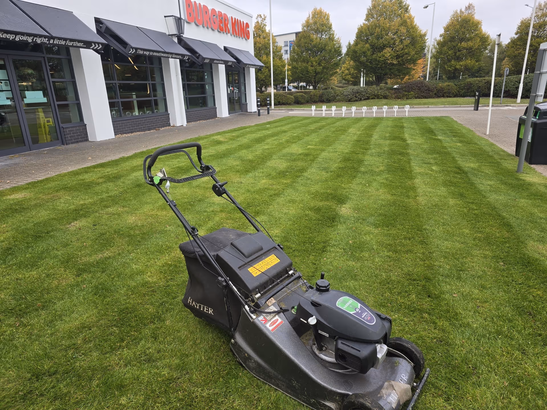Lawn mower on a striped green lawn in front of a Burger King restaurant on a cloudy day.
