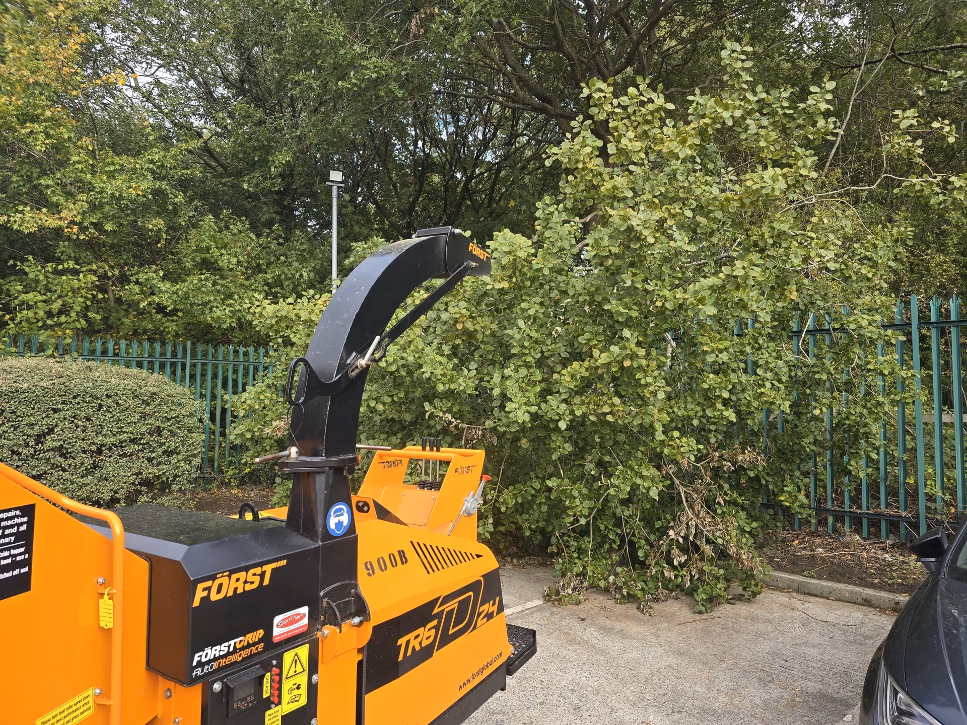 Orange Först TR6D24 wood chipper parked near green trees and a parked car on concrete.