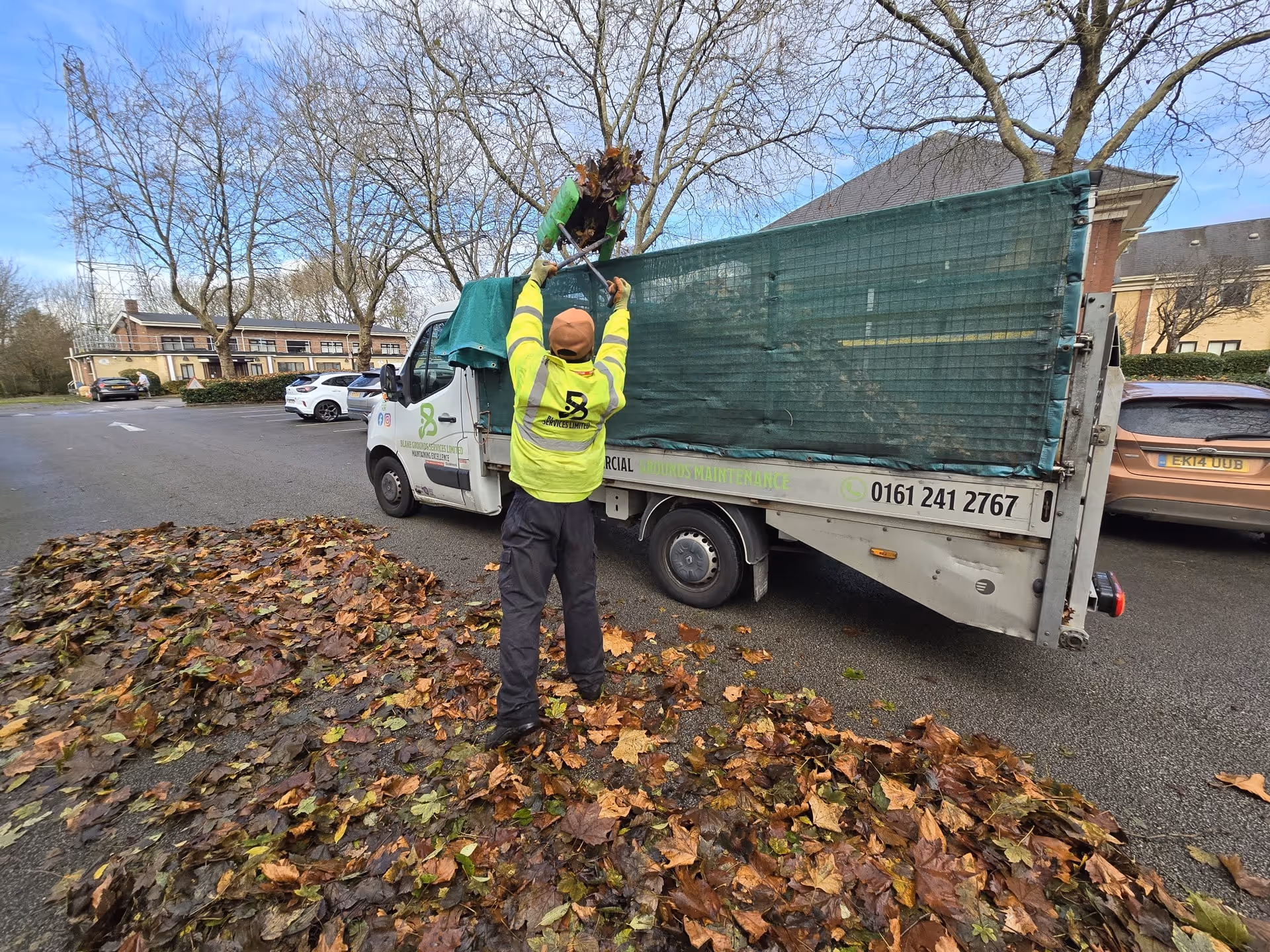 Worker in high-visibility jacket loads fallen leaves into a green-netted truck bed in parking lot.