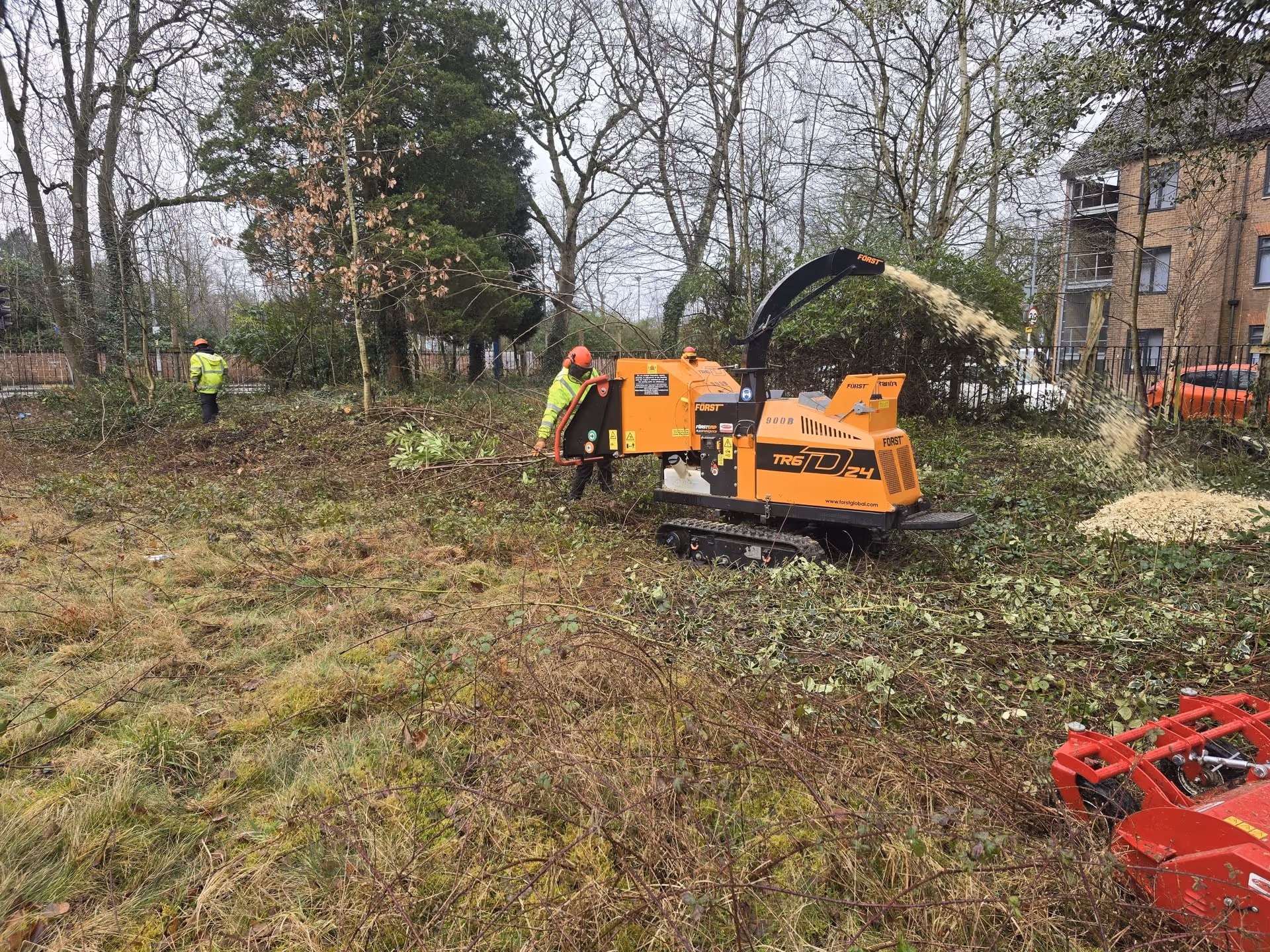 Workers in safety gear using a wood chipper to shred branches in a wooded urban area.