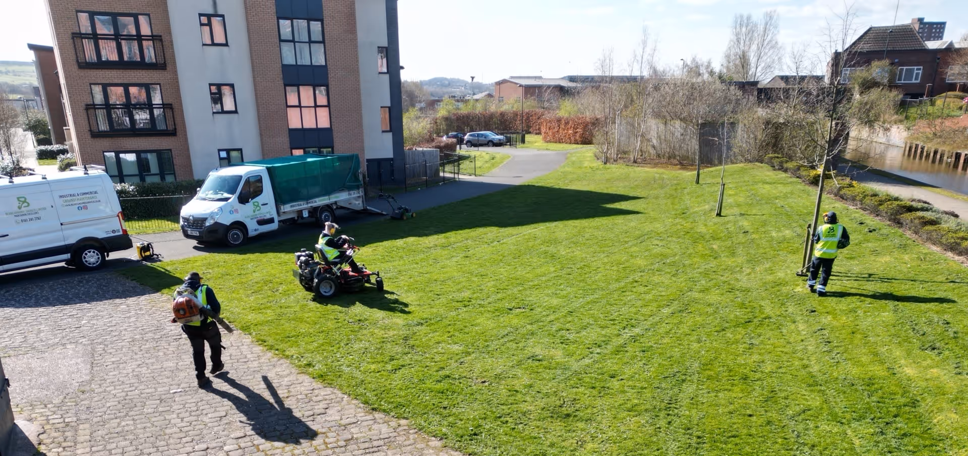 Three workers in yellow vests mowing a large green lawn near buildings on a sunny day.