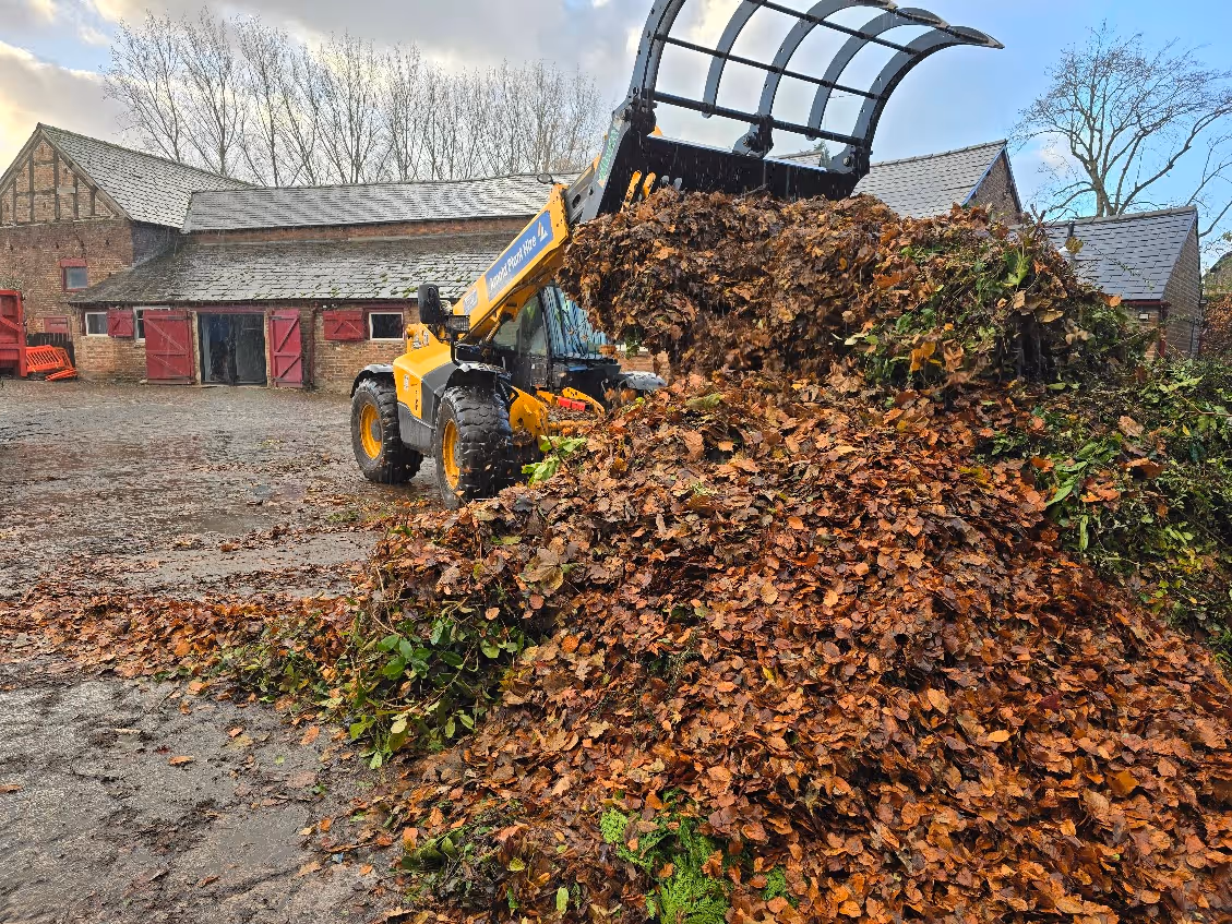 Yellow loader dumping a large pile of autumn leaves and green plant debris in a farmyard.