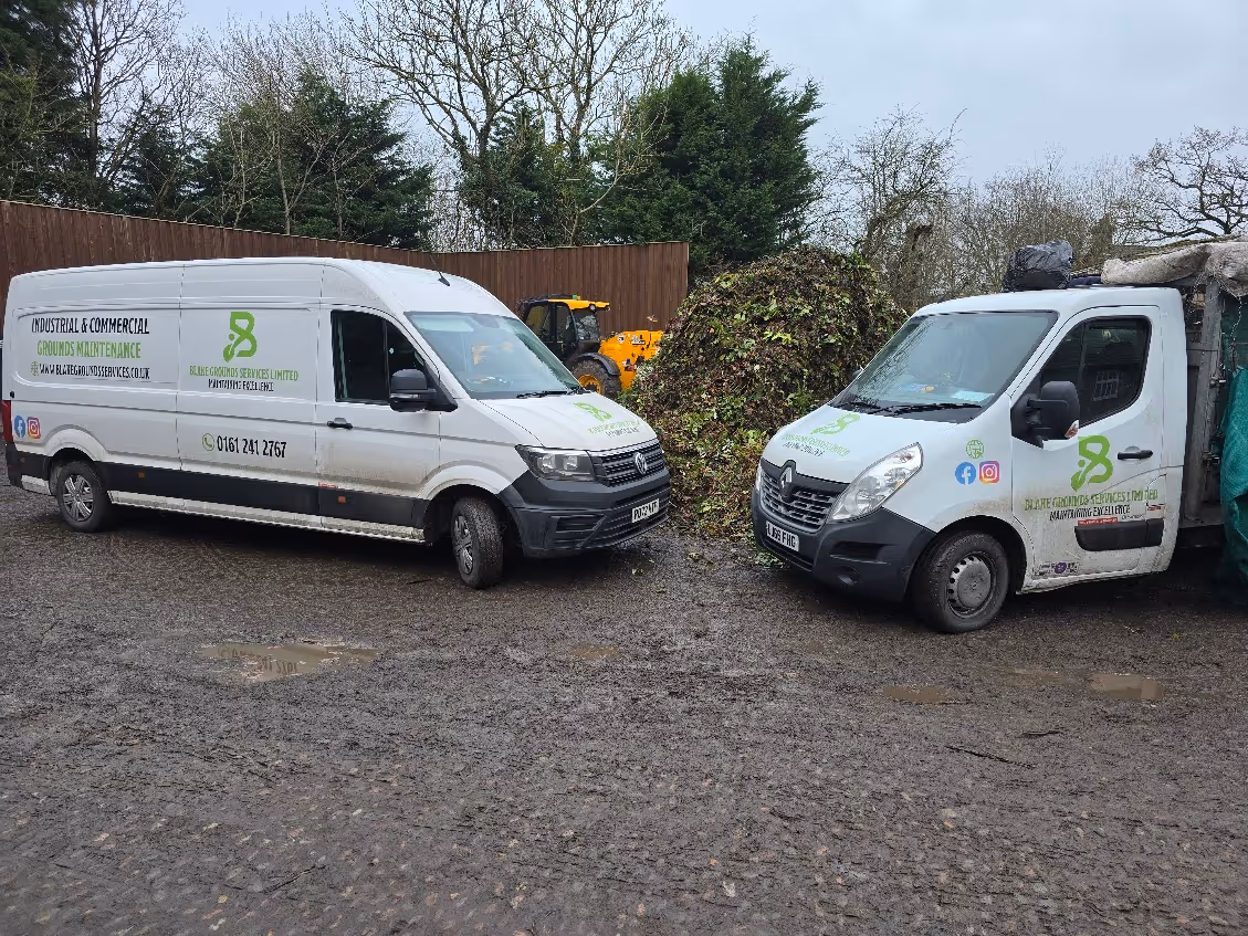 Two white commercial vans parked on gravel near a pile of vegetation and trees in background.