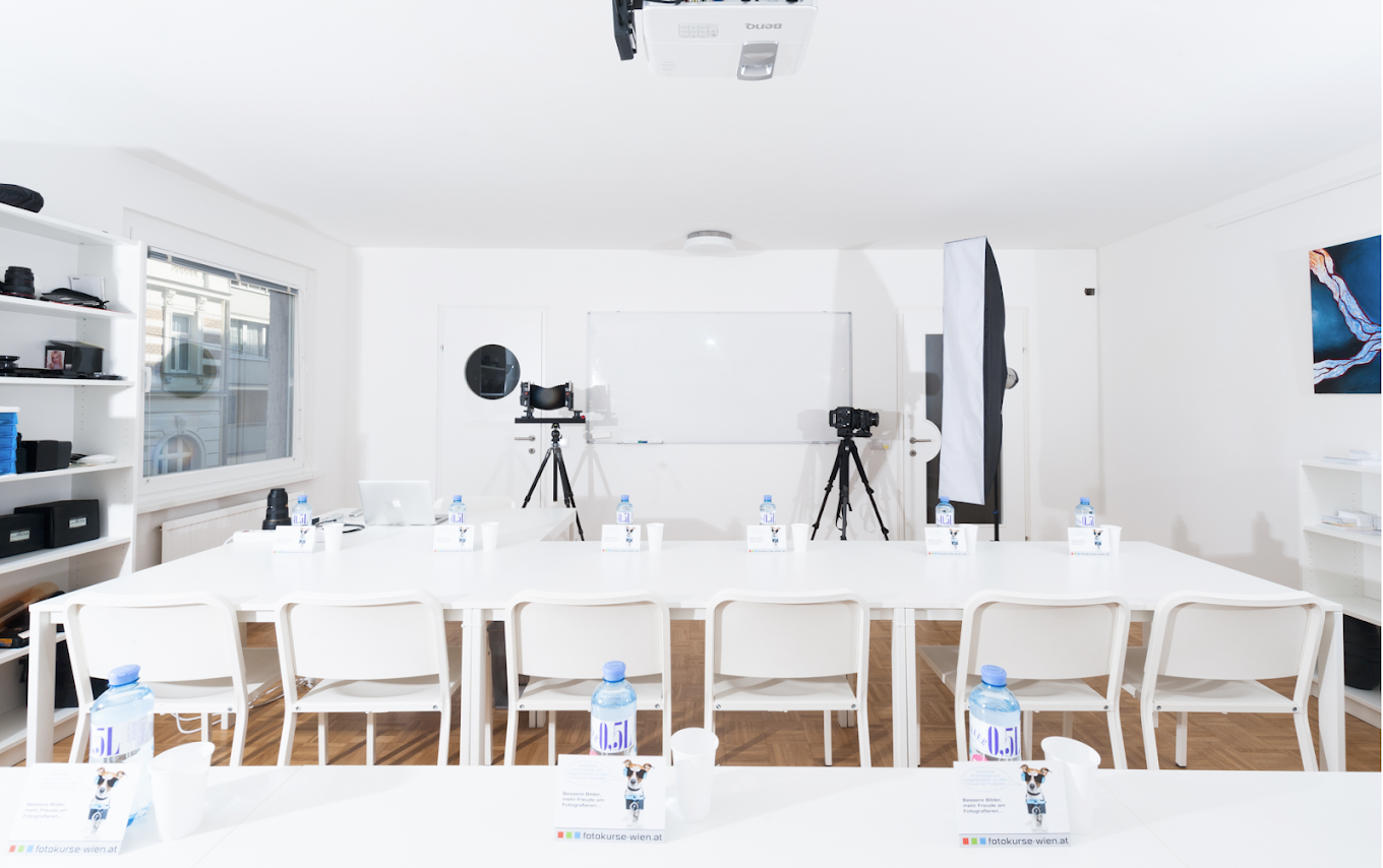 Helles Fotostudio mit weißen Tischen und Stühlen, Fotokameras auf Stativen und Whiteboard im Hintergrund.