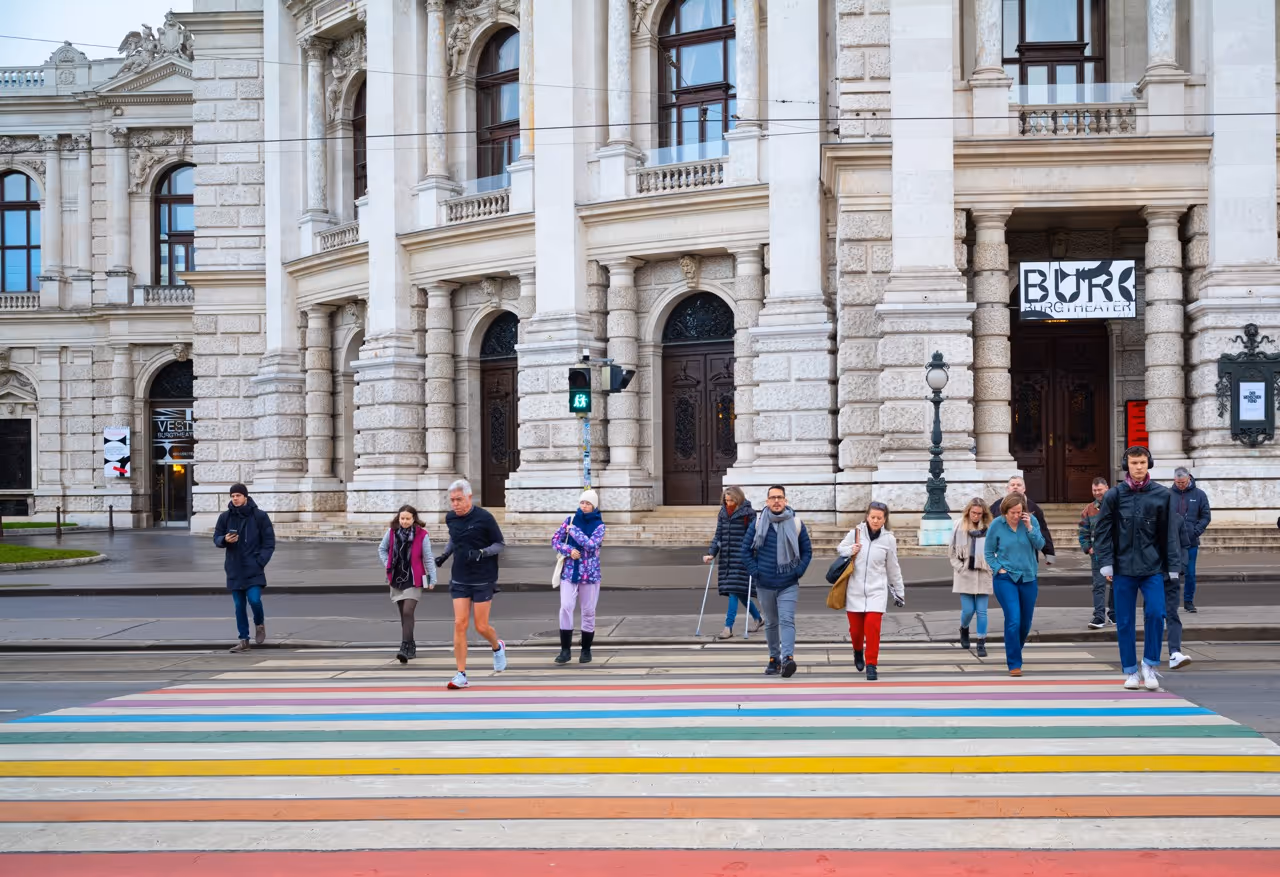 Menschen überqueren einen bunten Zebrastreifen vor einem historischen Gebäude in Wien.