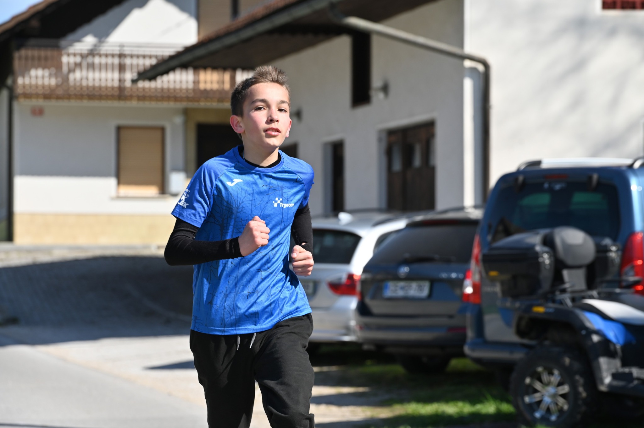 Fant otrok v modri športni majici in črnih hlačah teče po ulici v bivalnem naselju z avtomobili ob robu.