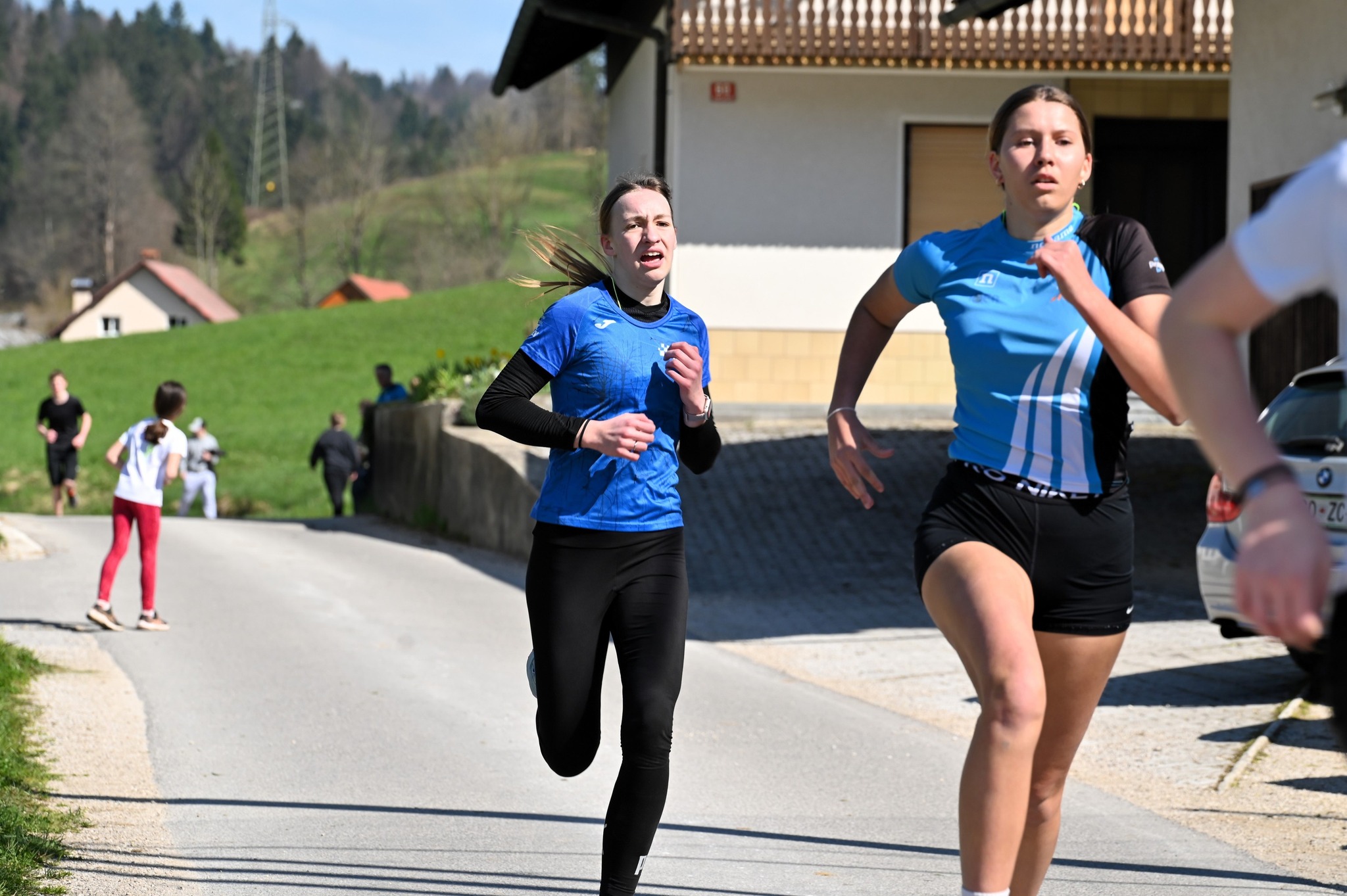 Tekačici na cesti v sončnem dnevu, z zelenim hribovitim ozadjem in nekaj stavbami.