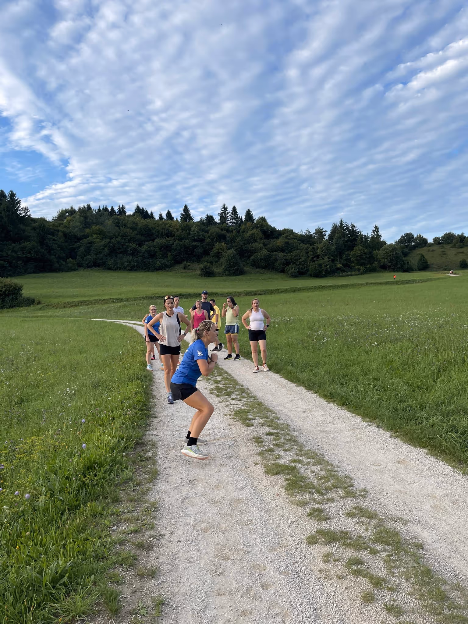 Skupina žensk, ki vadijo na makadamski poti v naravi ob oblačnem nebu.