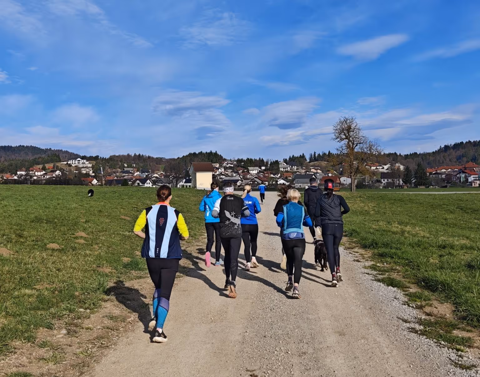 Skupina ljudi teče po makadamski poti skozi zeleno podeželje pod modrim nebom.