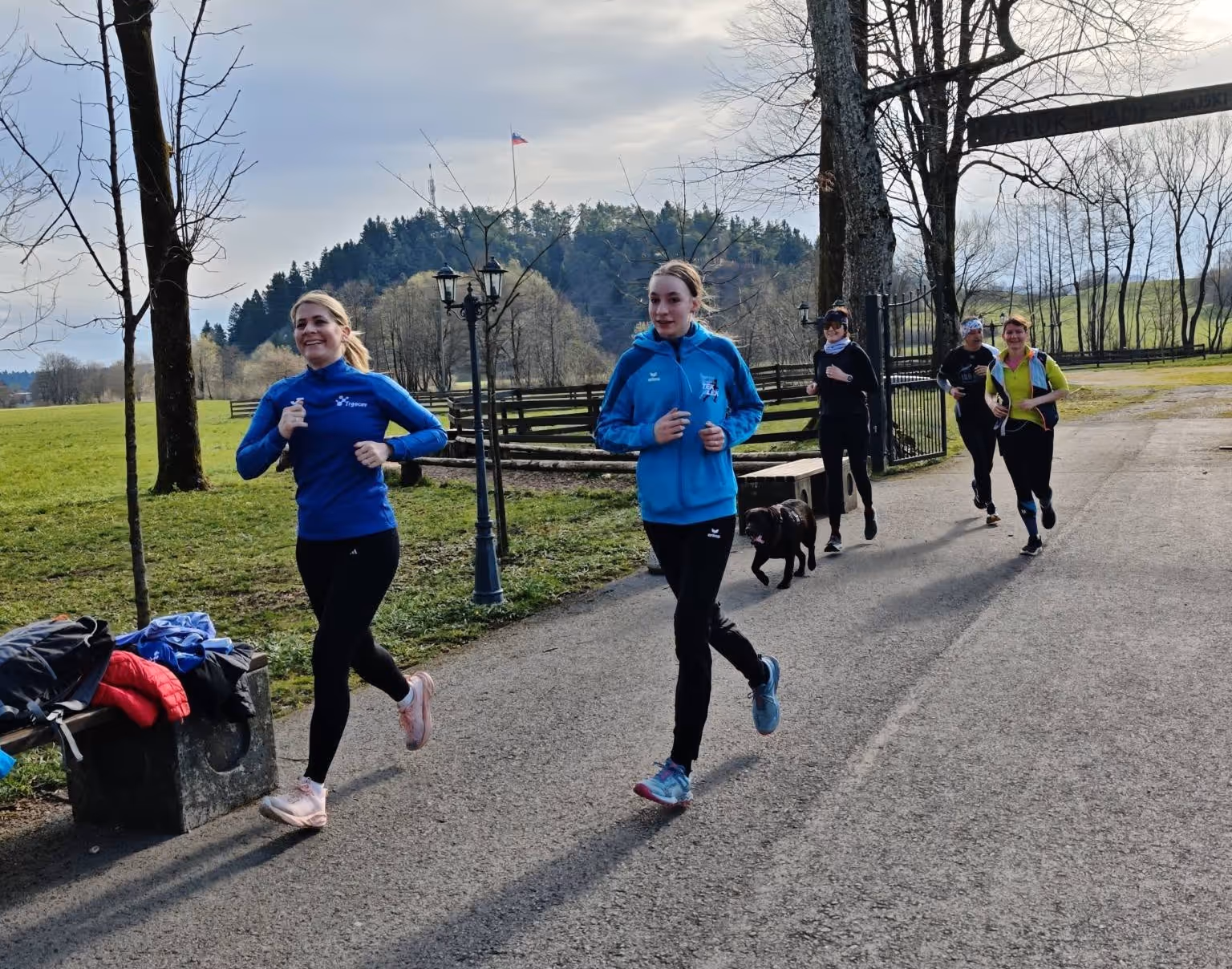 Pet žensk, ki tečejo po asfaltirani poti v podeželskem okolju, medtem ko teče tudi črn pes.