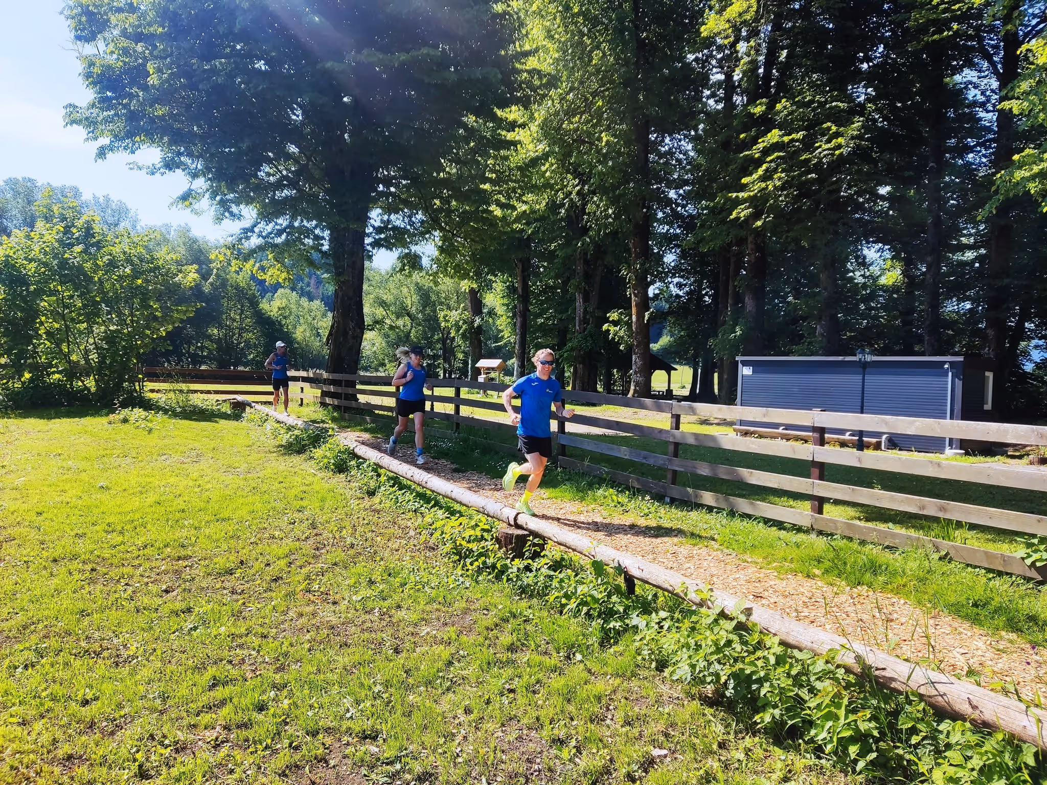 Trije ljudje tečejo po ozki gozdni poti ob leseni ogradi na sončen dan.