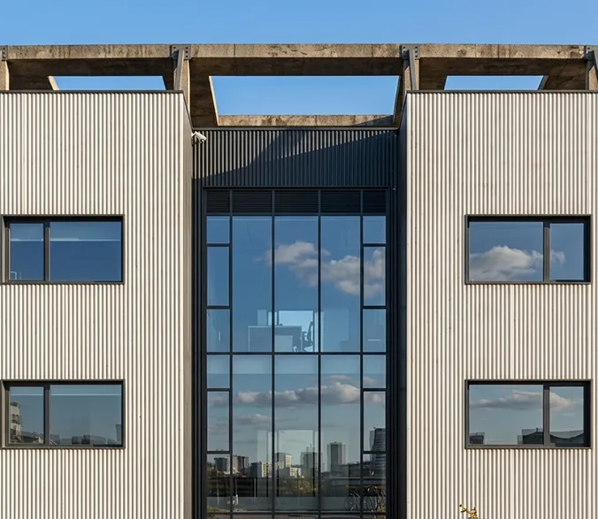 Modern building facade with large central glass windows reflecting clouds and surrounding cityscape.