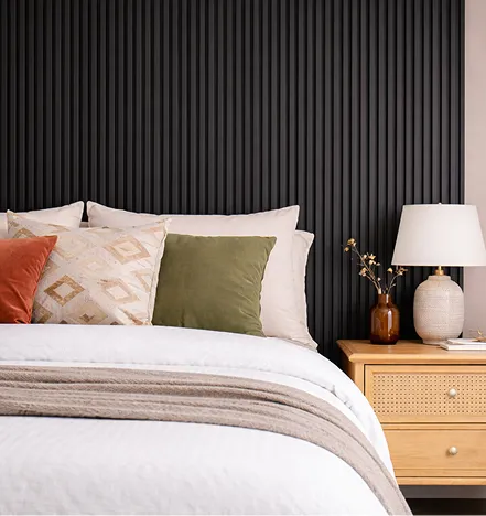 Close-up of a bed with white bedding, beige and green pillows, a wooden nightstand with a white lamp, and a brown vase against a black vertical slat wall.