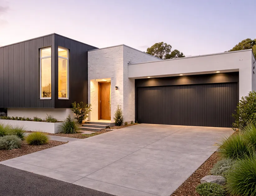 Modern single-story house with dark garage door, large window, wooden front door, and landscaped yard at dusk.