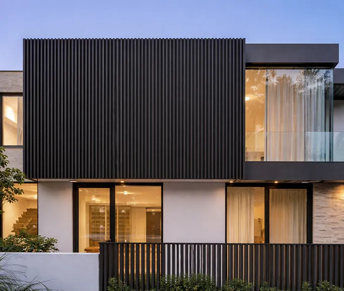 Modern two-story house exterior with vertical black paneling on upper floor, large windows, and illuminated interior at dusk.