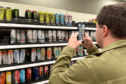 Man using a handheld device to photograph a shelf of Monster Energy and other energy drink cans