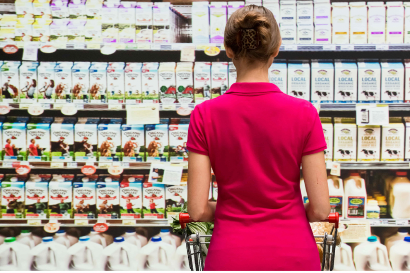 Shopper with a trolley facing a fully stocked dairy aisle of organic milk cartons and bottles